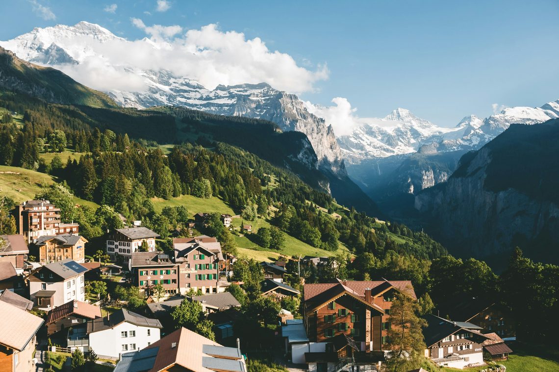 Un villaggio alpino svizzero su un pendio verde, con cime innevate sopra una valle profonda